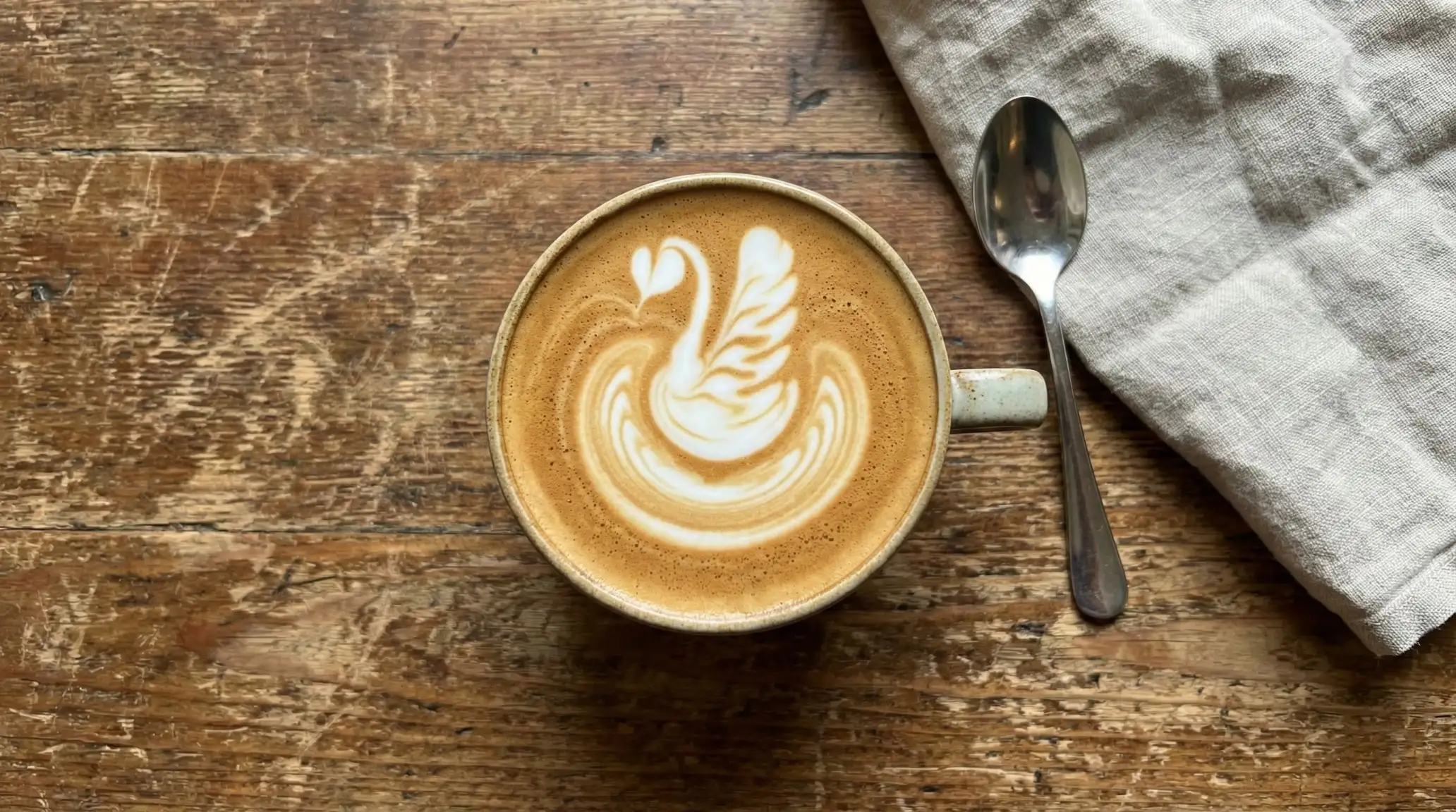 Fotografía cenital con móvil de un Flat White con arte latte de un cisne en una taza de cerámica sobre una mesa de madera rústica.